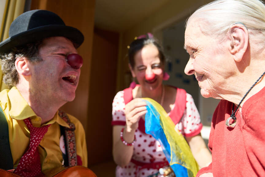 Ein männlicher Clown lacht eine ältere Dame an, die ihn zurück anlächelt.
