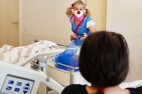 Ein weiblicher Clown schaut einer jungen Patientin, die im Krankenbett sitzt, in die Augen.