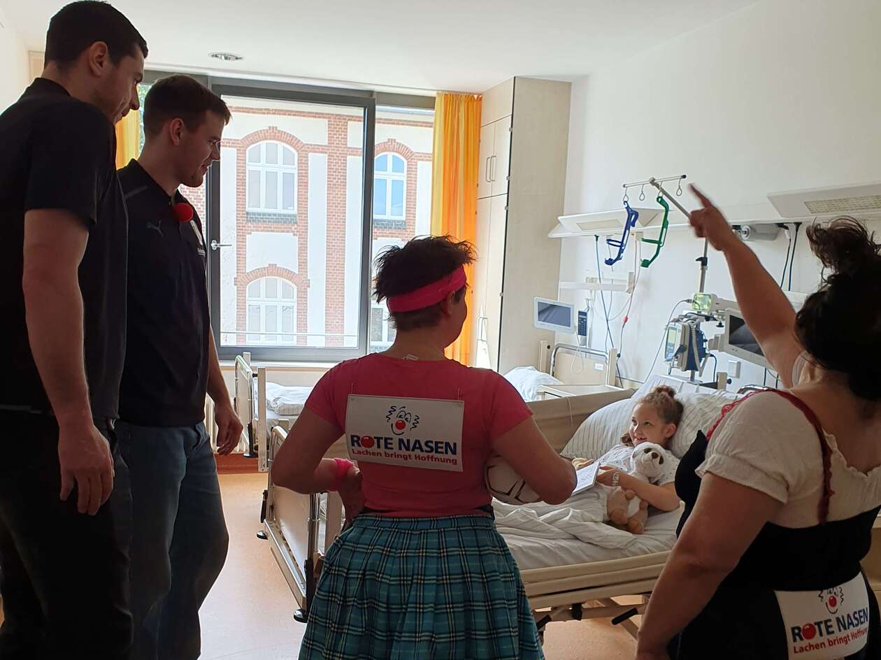 Zwei Handballspieler und zwei Clowns besuchen ein krankes Mädchen an ihrem Krankenhausbett. 