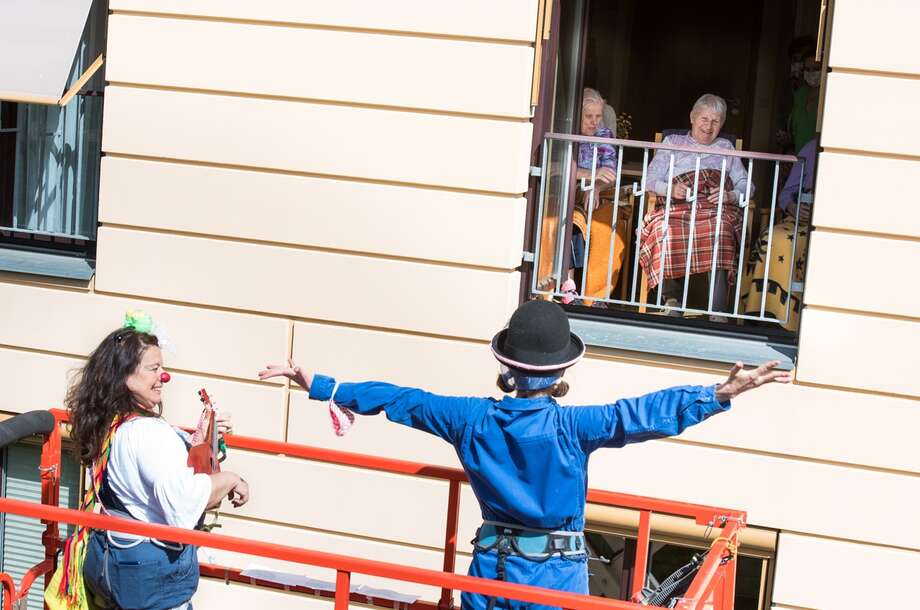 zwei weibliche Clowns seteh auf eienr hebebühne und singen drei älteren Damen im Rollstuhl in ihr fenster.