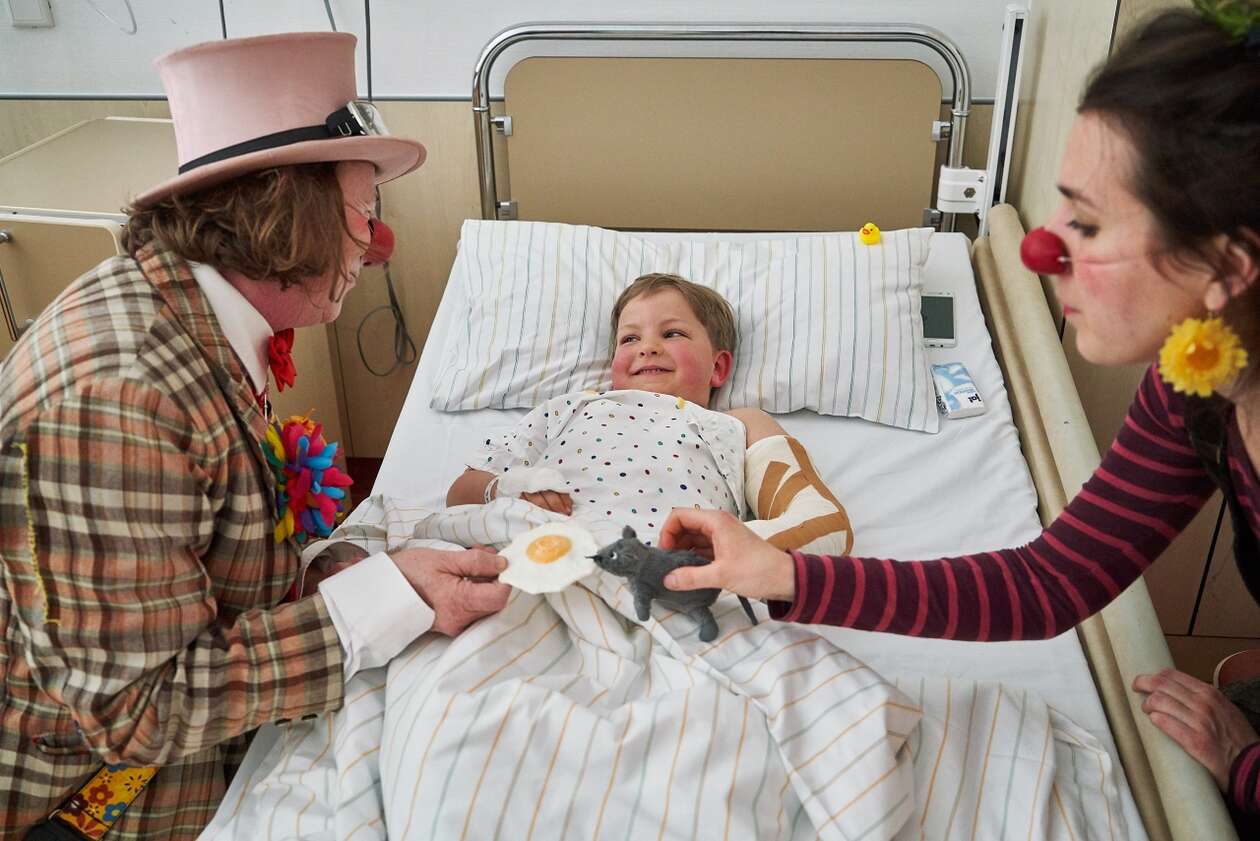 Die Clowns sietzen rechts und links von einem Kind am Bett und schauen auf es hinab 