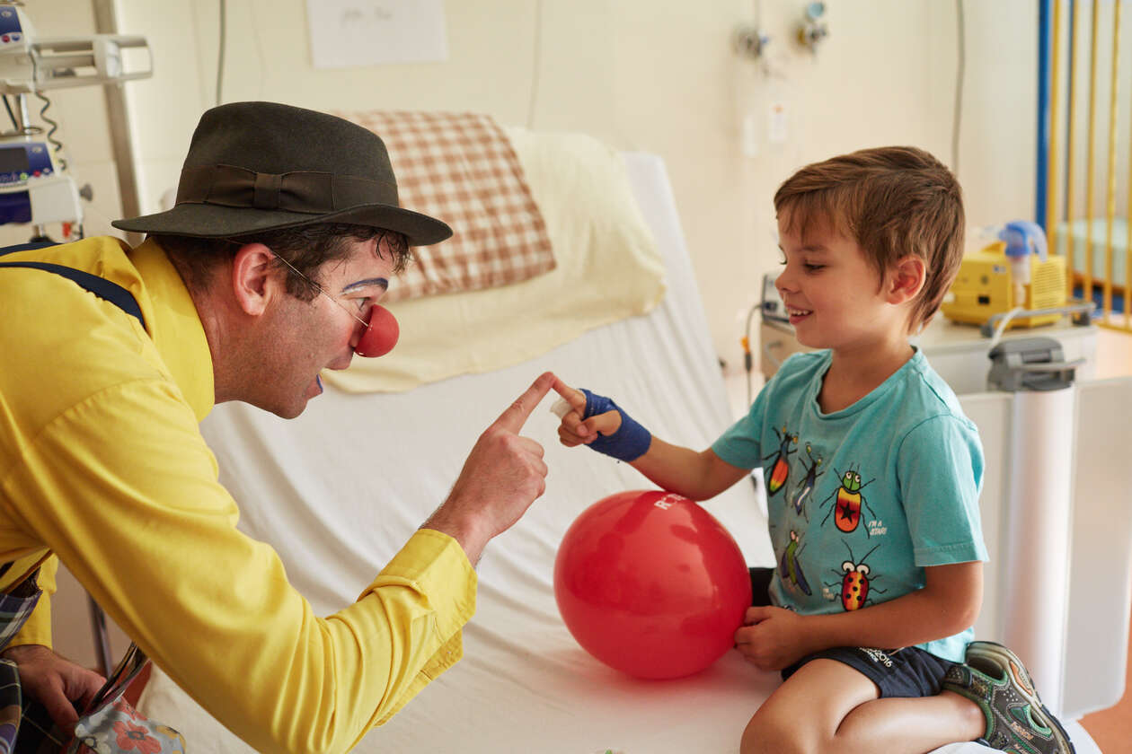  Der Klinikclown berührt den Zeigefinger eines kleines Jungen auf seinem Zimmer