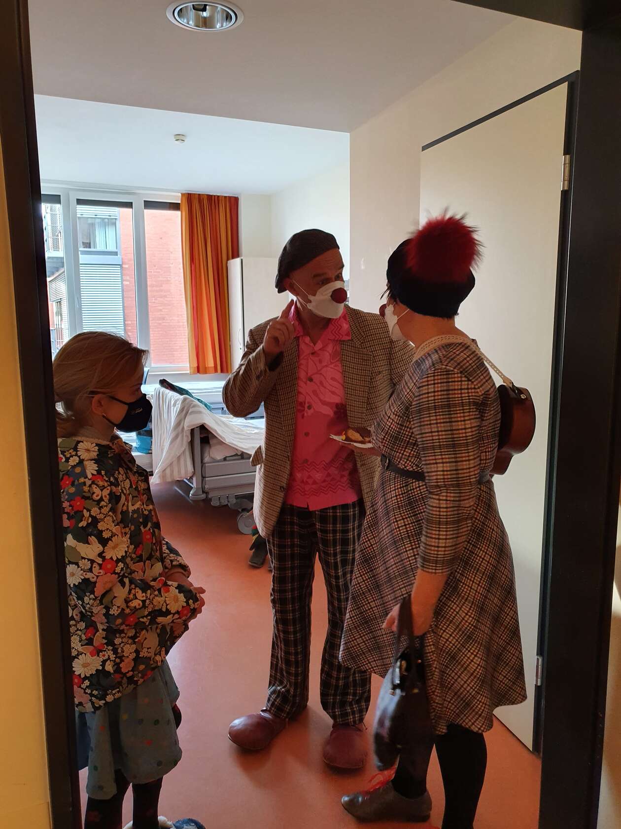 2 Clowns stehen im Zimmer mit einer Patientin und essen Kuchen