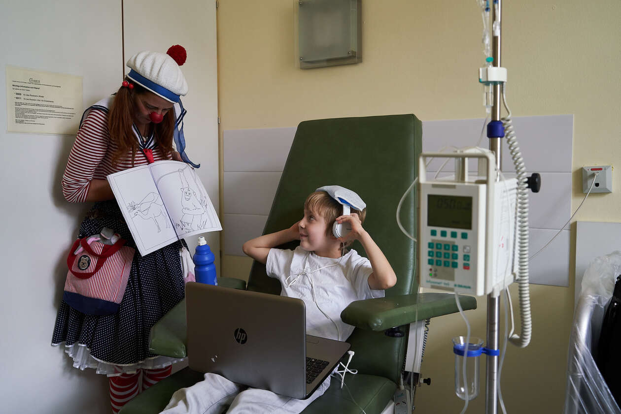 Ein Clown steht neben einem Kind was auf einem Stuhl vor einem Leptop sietzt 