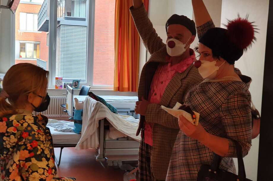 2 Clowns stehen im Zimmer einer Patientin und raten ein Ratespiel mit Kuchen in der Hand