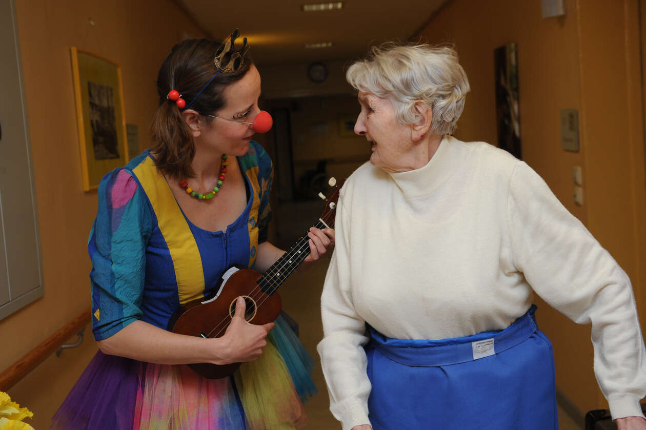 Ein Clown mit Krone und Ukulele schaut einer älteren Dame in die Augen und beide lächeln