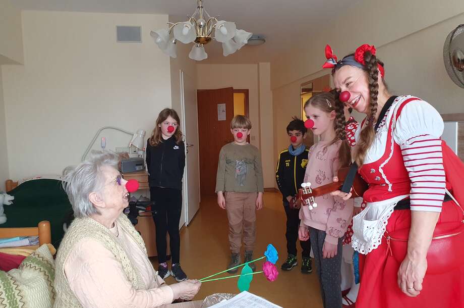 Ein Clown und vier Kinder mit roten Nasen stehen in einem Zimmer in einer Pflegeeinrichtung. Sie schauen eine ältere Dame an, die ebenso eine rote Nase auf hat und sie anlacht 