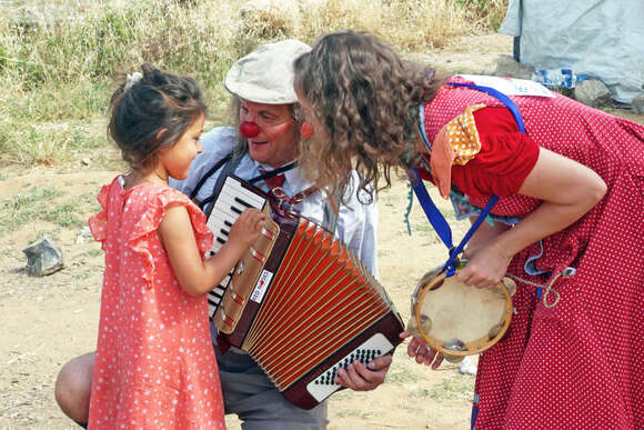 ROTE NASEN Clown Luciana im Flüchtlingscamp in Lesbos beugt sich mit roter Nase zu einem geflüchteten Mädchen hinunter. Ein anderer Klinikclown hat ein Akkordeon in der Hand. Im Rahmen des Programms Emergency Smile.