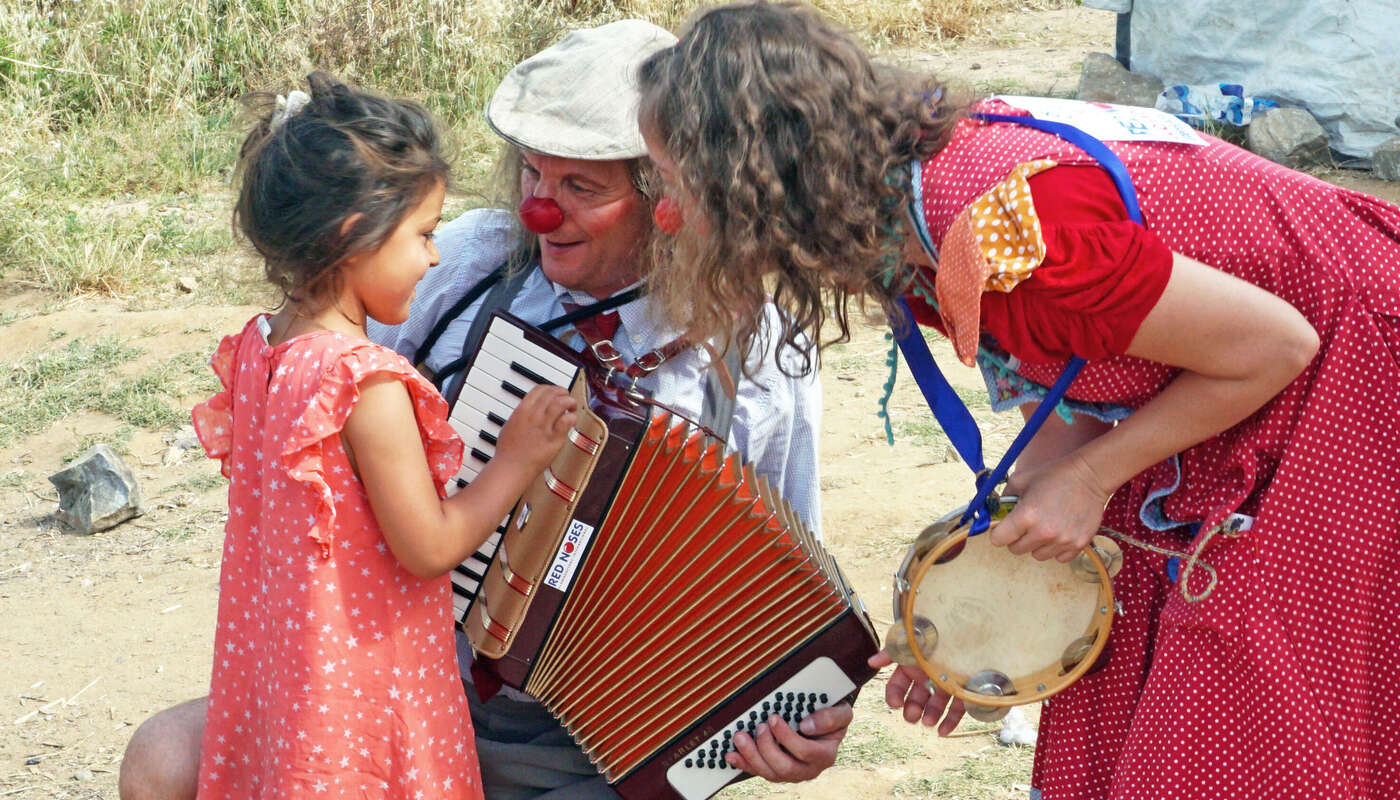 ROTE NASEN Clown Luciana im Flüchtlingscamp in Lesbos beugt sich mit roter Nase zu einem geflüchteten Mädchen hinunter. Ein anderer Klinikclown hat ein Akkordeon in der Hand. Im Rahmen des Programms Emergency Smile.