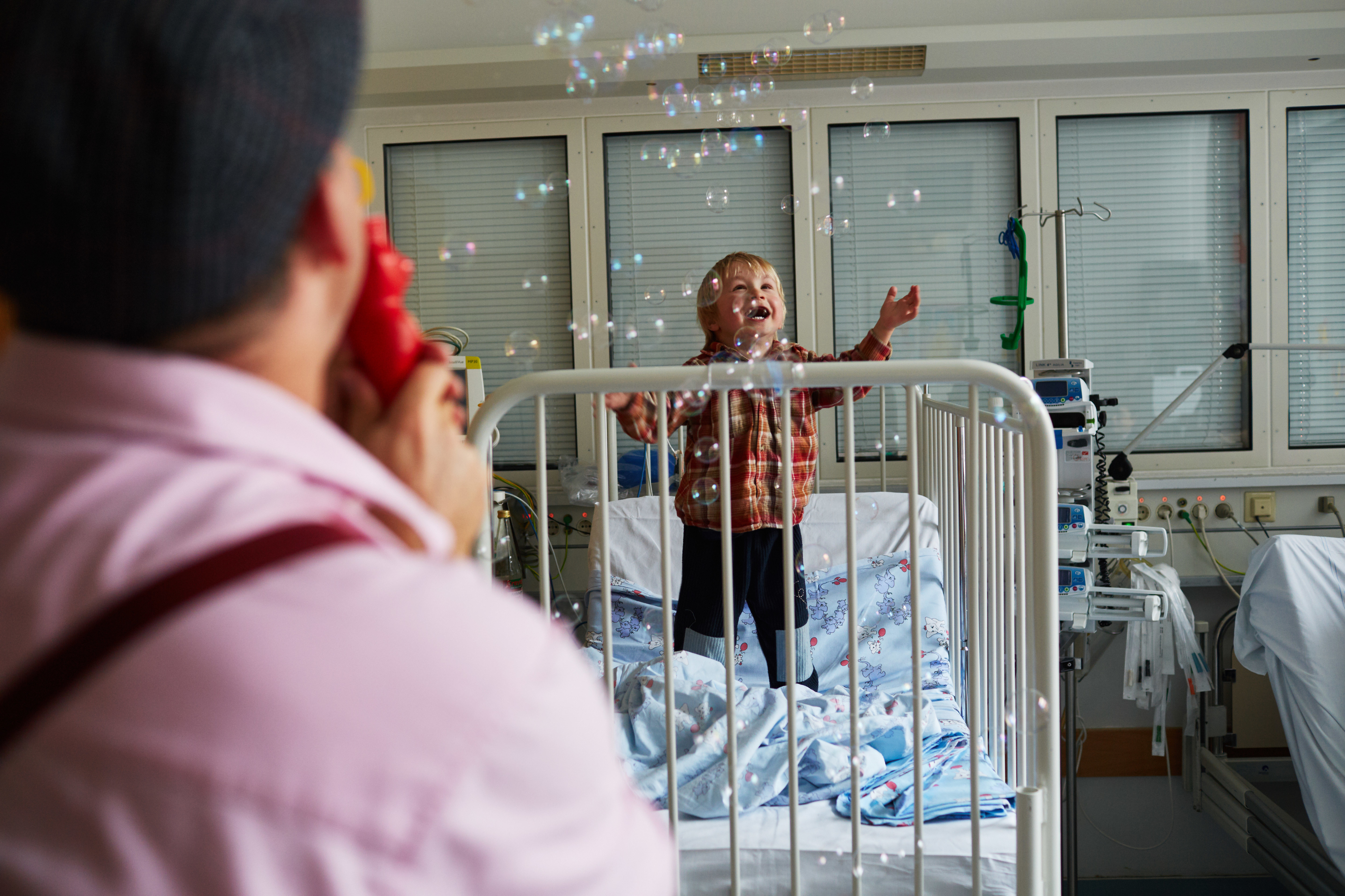 Ein Clown pustet ganz viele kleine Seifenblasen in ein Krankenzimmer von einem kleinem Jungen, der versucht nach ihnen zu greifen 